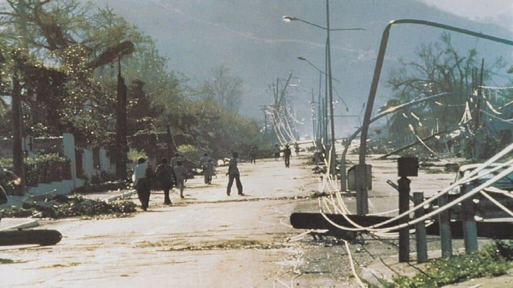 Photo of Hope Road Kingston in the aftermath of Hurrica Gilbert showing fallen light posts and wires, windswept trees, and people walking around surveying the damage.